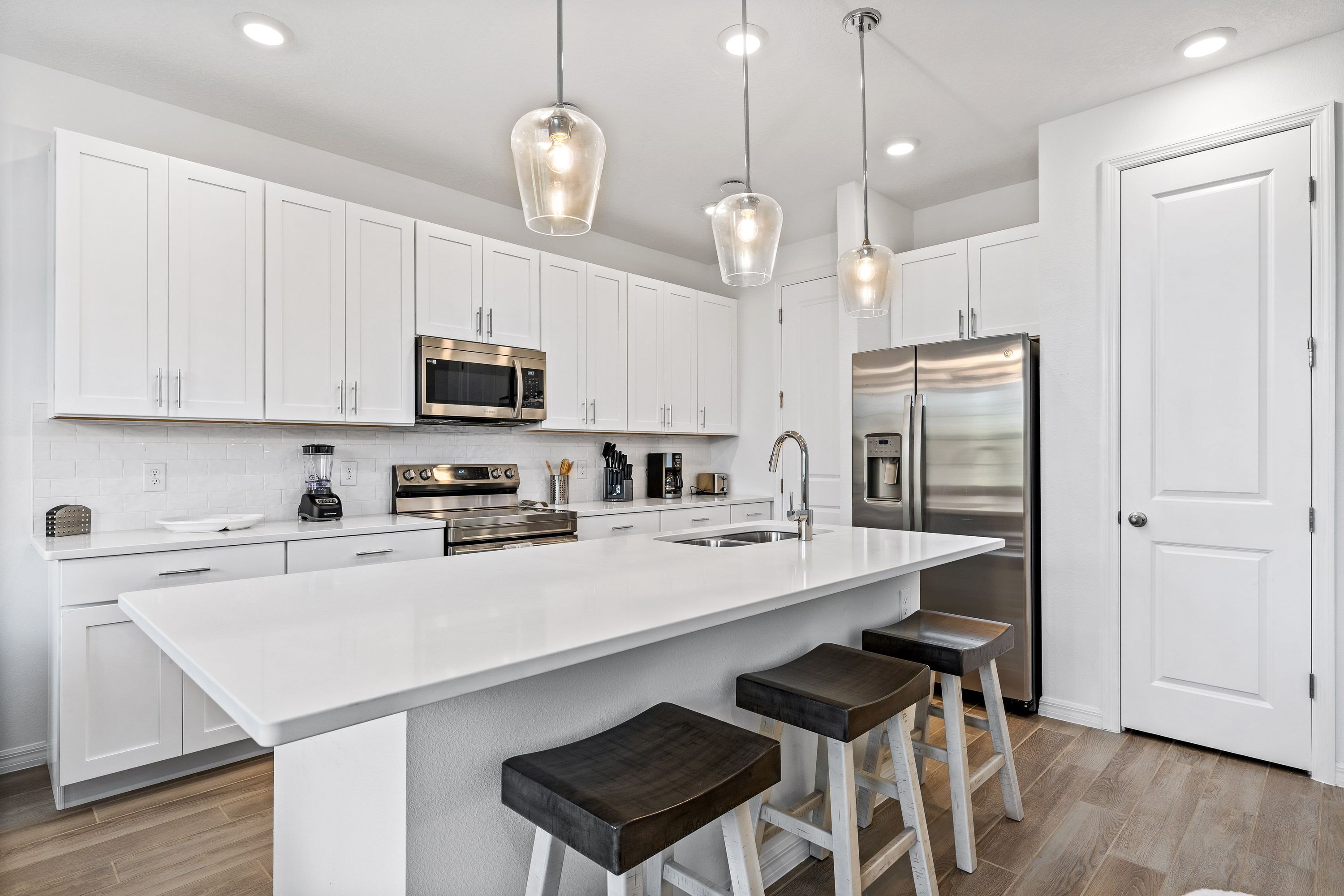 A modern kitchen with white cabinetry, stainless steel appliances, a large island with stools, and pendant lighting.