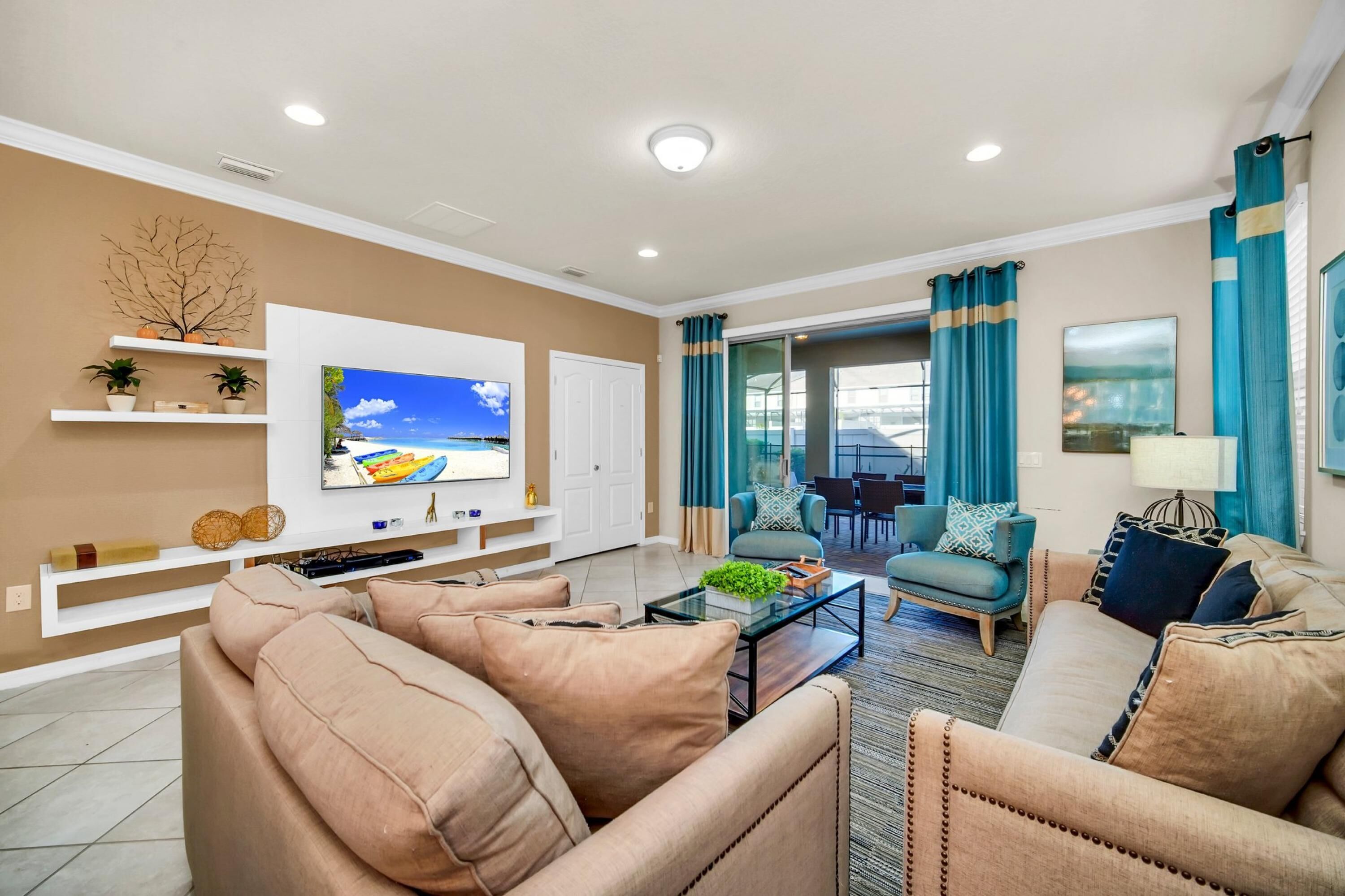 A modern living room with beige sofas, a glass coffee table, and a flat-screen TV on a white wall. There are blue curtains, a dining area, and a view of the outdoors.
