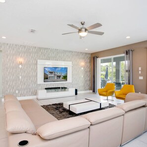 A modern living room with a large sofa, a coffee table, and a TV mounted on the wall. There is a ceiling fan and a window with curtains.