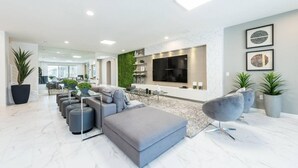 A modern living room with a grey sofa, ottoman, and a TV mounted on the wall. There are potted plants and a dining area in the background. - Unique Masterpiece15 Suites Sleeps 30 Son101 (Kissimmee)