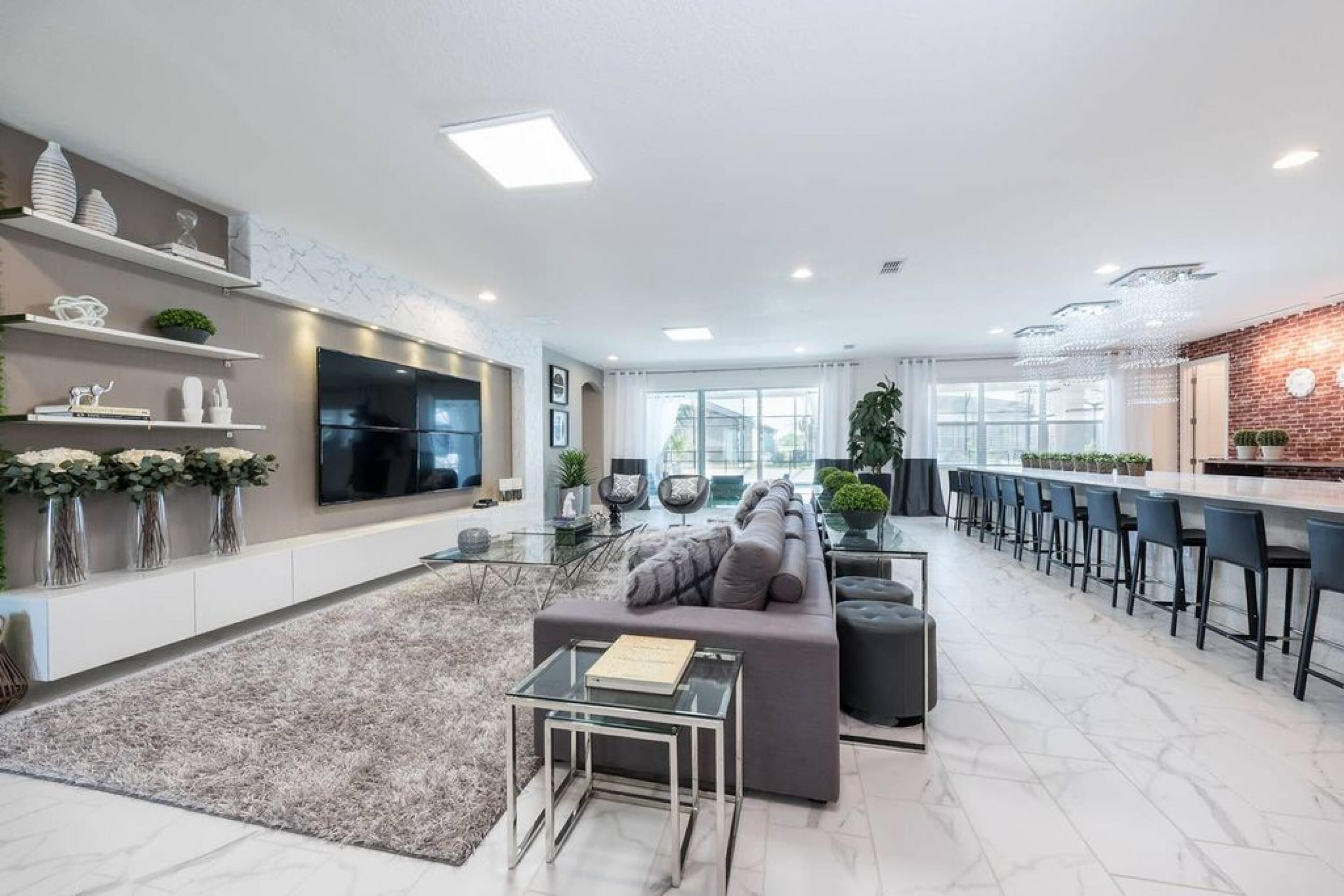 A modern living room with a grey sofa, a glass coffee table, and a TV mounted on the wall. There is a kitchen area with a bar and stools, and a large window providing natural light.