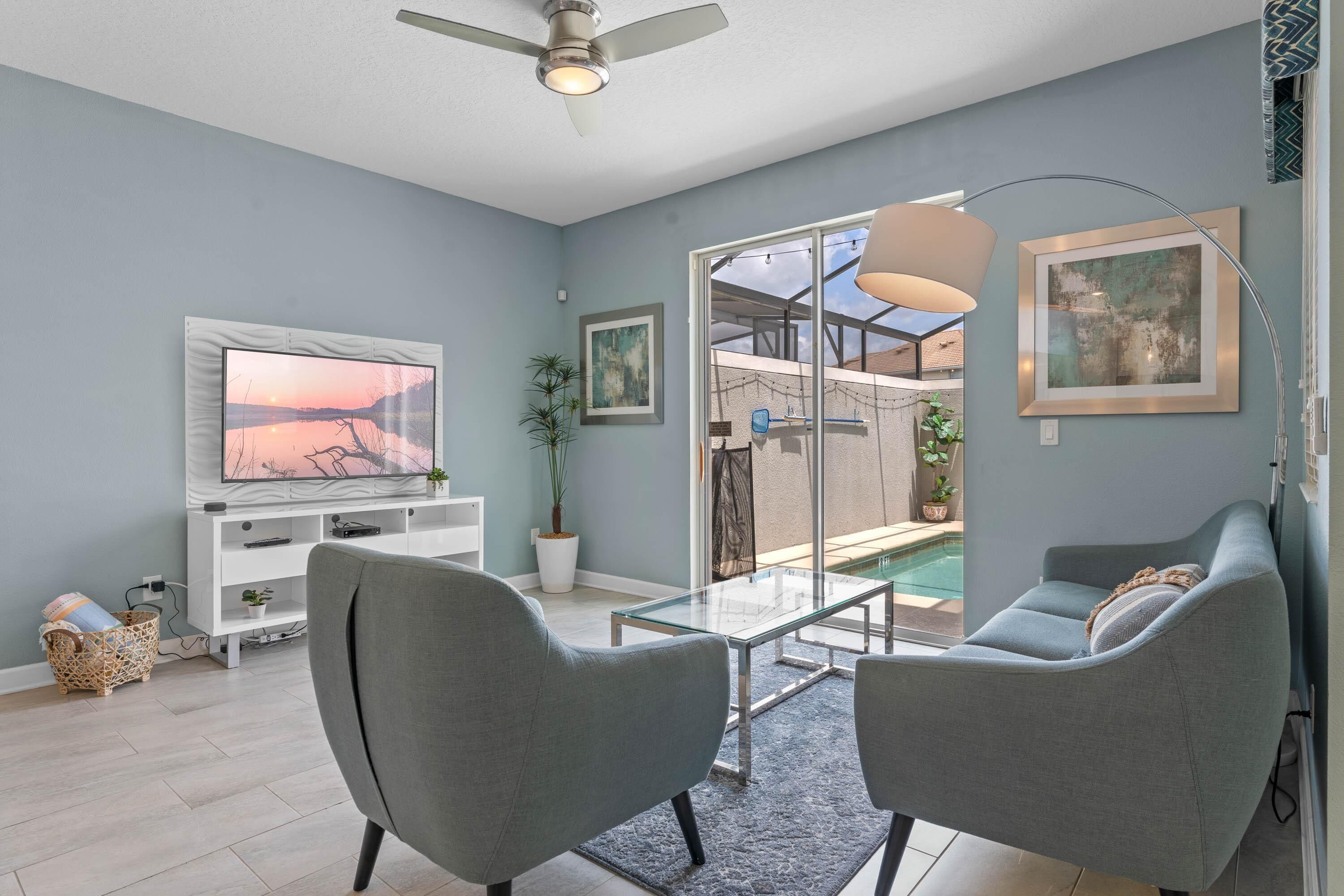 A modern living room with a flat-screen TV, grey sofas, a glass coffee table, and a ceiling fan.