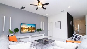 A modern living room with a flat-screen TV, a glass coffee table, and a ceiling fan.