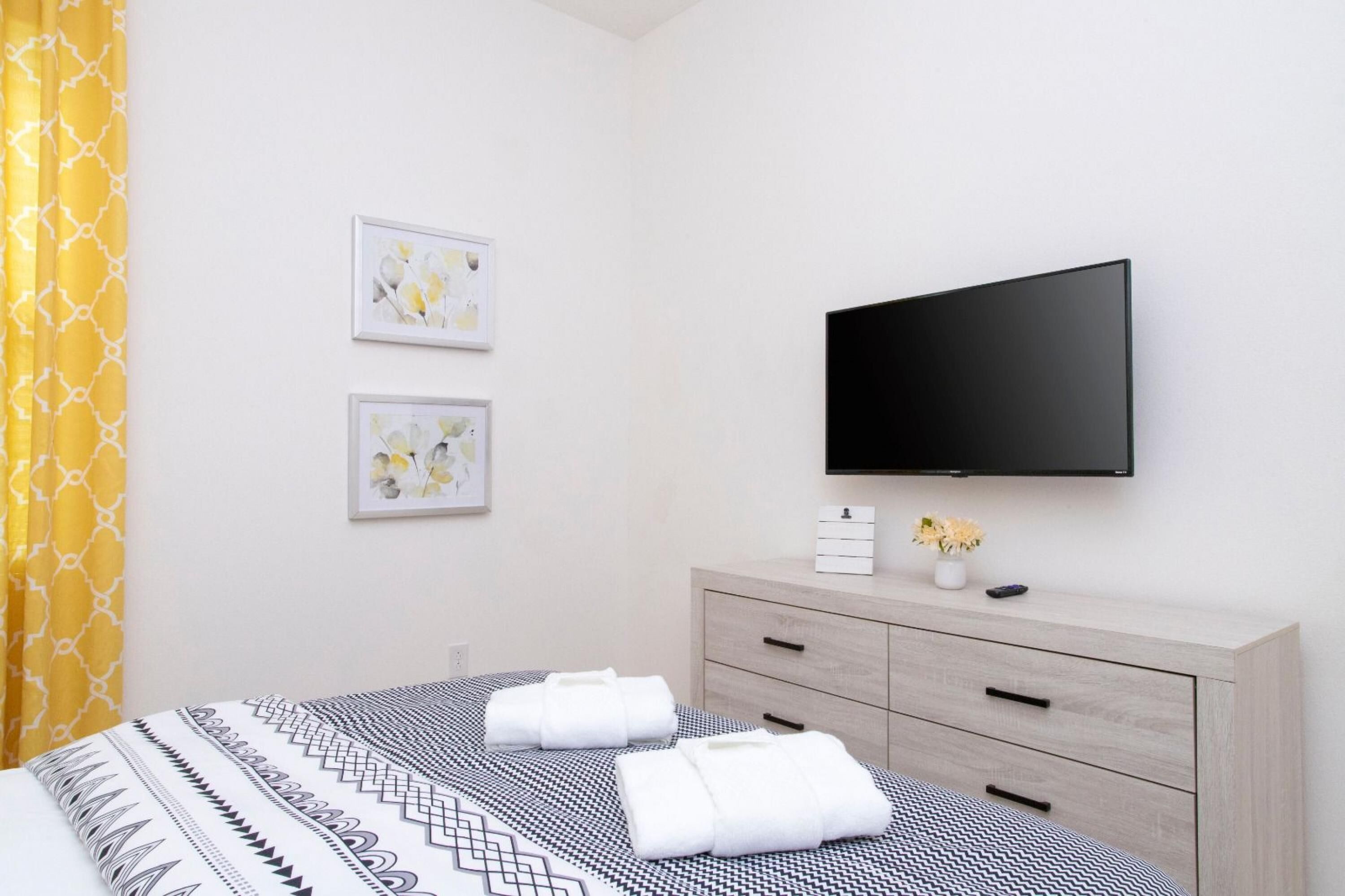 A hotel room with a bed, a TV mounted on the wall, a bedside table with a vase of flowers, and two framed pictures on the wall.