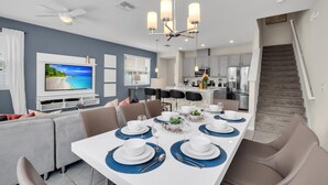 A modern dining area with a large table set for six, a ceiling fan, and a kitchen in the background. - Comfy House at Compass Bay Cb102 (Kissimmee)
