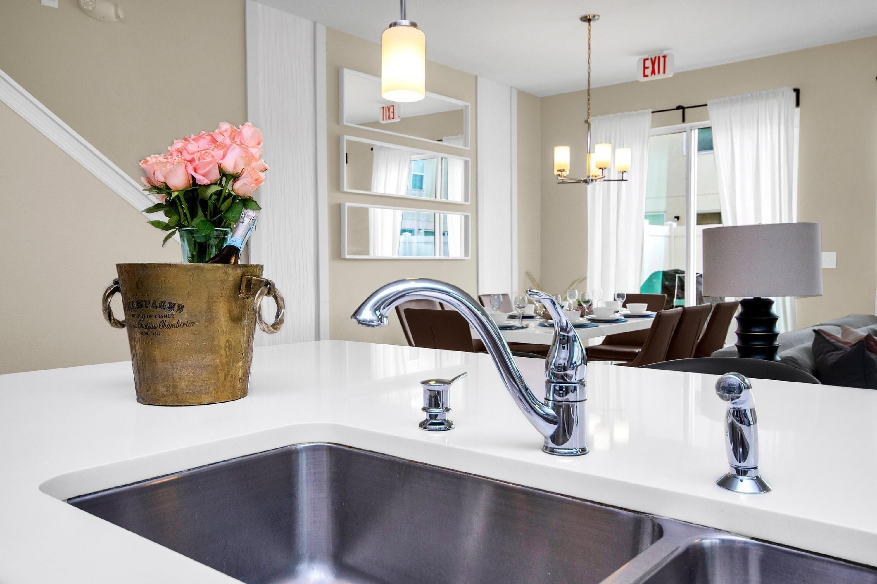 A modern kitchen with a stainless steel sink, a brass bucket with flowers, and a dining area in the background.