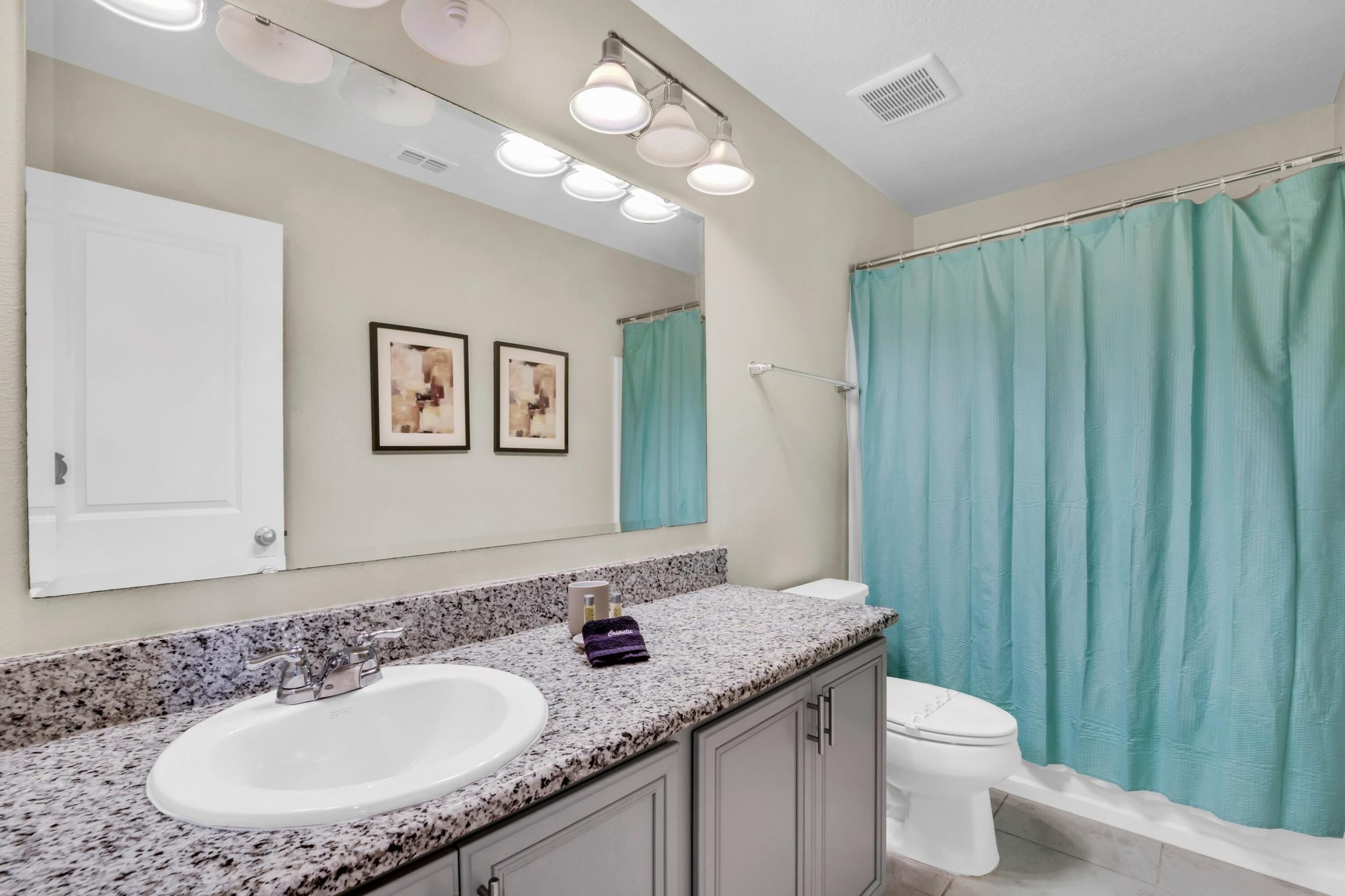 A bathroom with a granite countertop sink, a white toilet, a teal shower curtain, and a mirror with three lights.