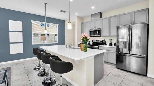 A modern kitchen with grey cabinets, a stainless steel refrigerator, a white island with bar stools, and pendant lighting. - Comfy House at Compass Bay Cb102 (Kissimmee)