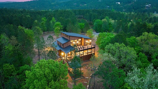 A modern house with a large deck and multiple windows, surrounded by dense forest. - Stars & Streams - Ev Outlet, Pool Table,Shuffleboard, Hot Tub, Arcade Console (Broken Bow)