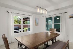 A dining area with a wooden table and chairs, large windows with curtains, and a hanging light fixture.