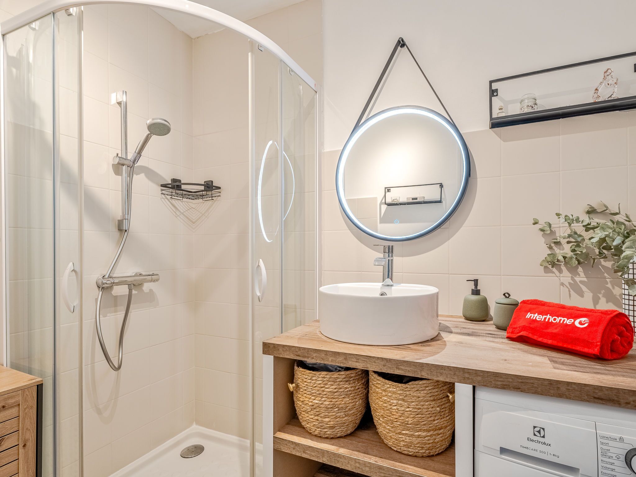 A modern bathroom with a glass-enclosed shower, a round mirror with a light, a white sink, and a wooden vanity.