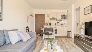 A modern living room with a grey sofa, a dining table with chairs, a kitchen area with white cabinets, and a flat-screen TV. - Le Manon by Interhome (Dinard)