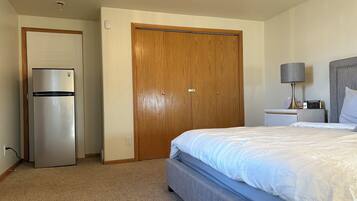 A hotel room with a bed, a wooden wardrobe, a refrigerator, a bedside table with a lamp, and a digital clock.