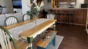 Ein Essbereich mit einem Holztisch, Stühlen und einer Blumenvase. Im Hintergrund ist eine Küche erkennbar.