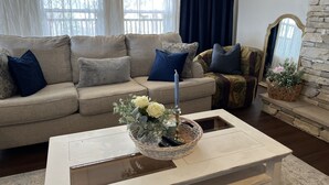 Ein Wohnzimmer mit einem beigen Sofa, blauen Kissen, einem weißen Couchtisch mit einer Blumenkorb-Dekoration, einer Kerze und einem Steinkamin.