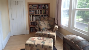 A cozy room with a wooden floor, a bookshelf, a patterned armchair, a window with a view of trees, and a door.