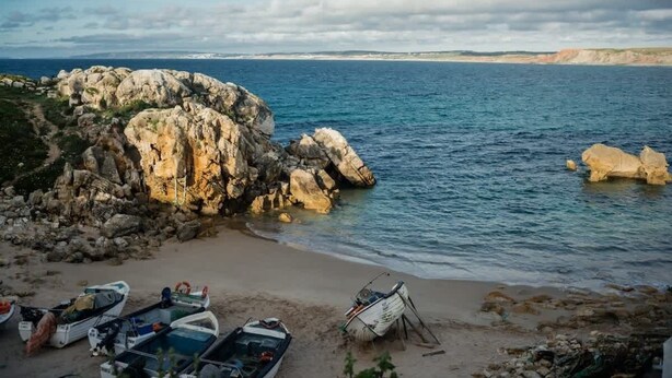 A coastal scene with rocky formations, calm sea, and boats on the shore.