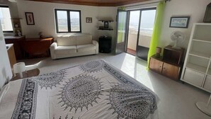 A bedroom with a bed, a sofa, a desk, a chair, a small table, a cabinet, and a sliding glass door leading to a balcony.
