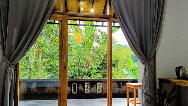 Una habitación con puerta de vidrio enmarcada en madera que da a un balcón con vistas a abundante vegetación.