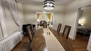 Ein Essbereich mit einem langen Holztisch, Stühlen und einem Teppich in der Mitte. Links befindet sich eine Heizung, im angrenzenden Raum ein Bett.
