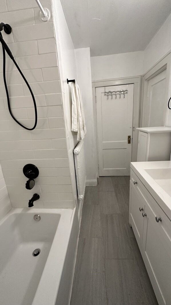 Une salle de bain avec une baignoire, une douche et des toilettes. On y trouve un porte-serviettes sur lequel sont suspendues des serviettes. Les murs sont recouverts de carreaux, et une porte donne accès à une autre pièce.