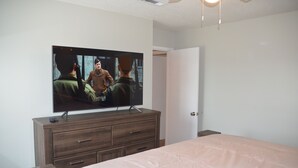 Un dormitorio con un televisor de pantalla plana grande montado en un aparador de madera, una cama con una colcha de color claro y una puerta blanca al fondo.