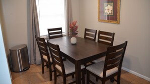 Un comedor con una mesa de madera y seis sillas, un cubo de basura y una ventana con cortinas.