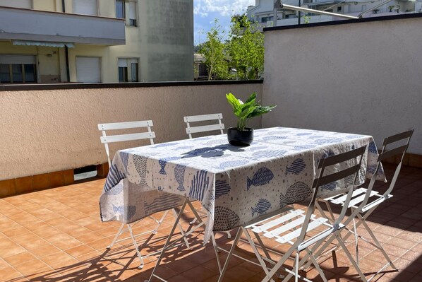 Een klein terras buiten, met een tafel bedekt met een blauw-wit geruit tafelkleed, omringd door witte stoelen.