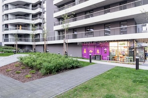 Un edificio moderno de varios pisos con balcones, una cafetería en la planta baja y un área ajardinada con arbustos y un camino pavimentado.