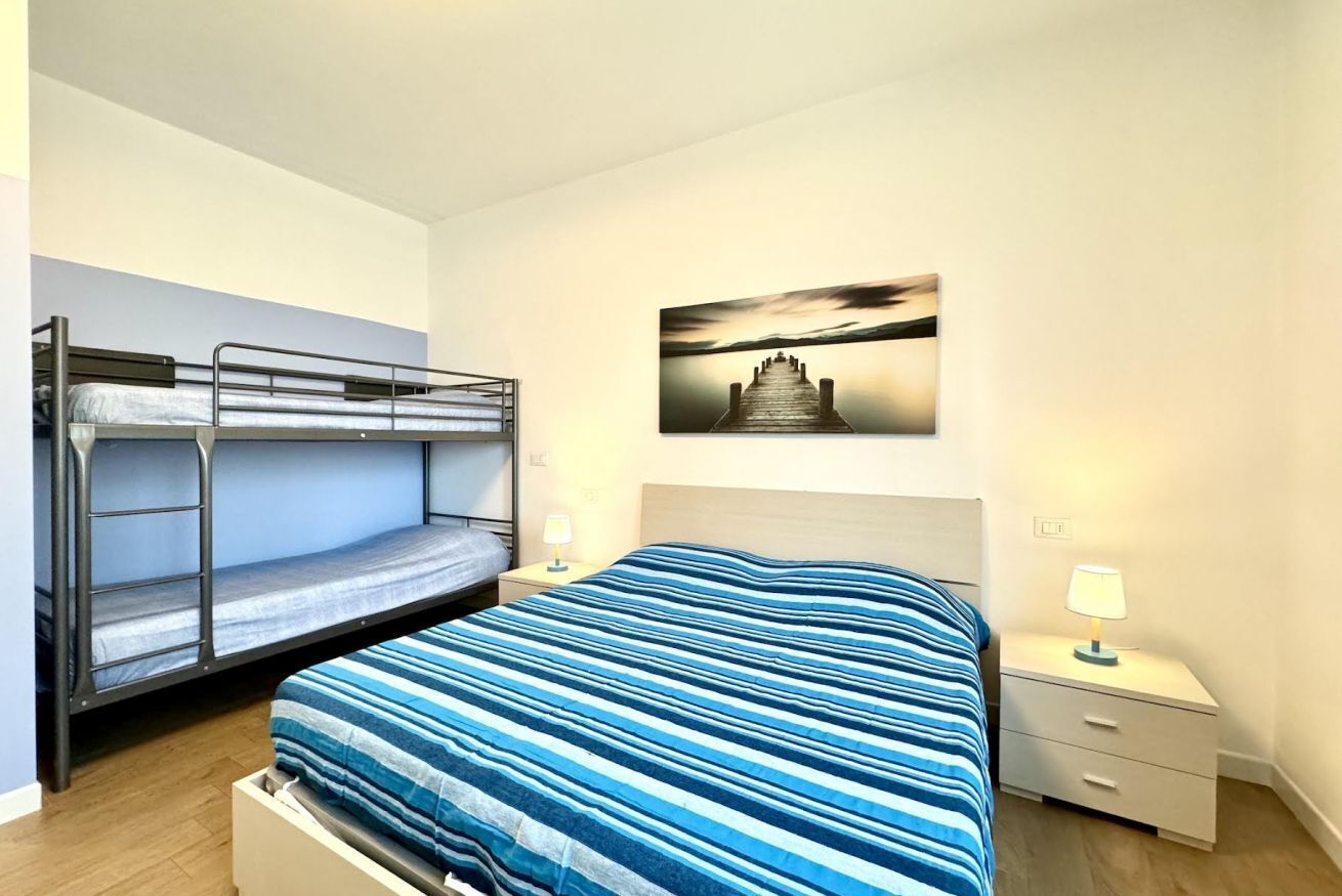 A bedroom with bunk beds, a single bed with a striped blue and white bedspread, a nightstand with a lamp, and a painting of a pier on the wall.