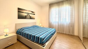 Une chambre à coucher avec un lit, une table de chevet, une lampe, un tableau au mur et une fenêtre avec des rideaux.