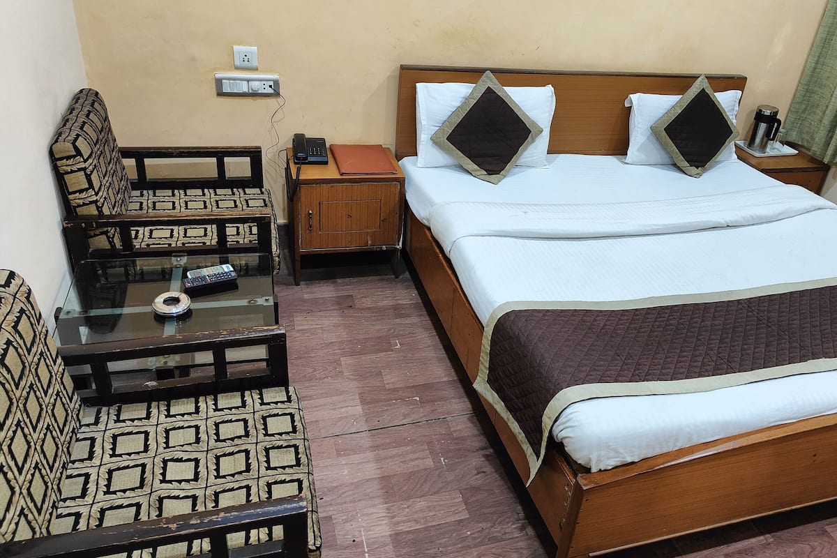 Una habitación de hotel con una cama, una mesita de noche con un teléfono, una pequeña mesa con un control remoto, un sillón estampado y una cortina verde.