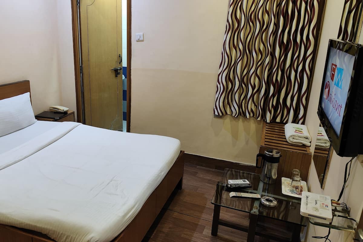 Habitación de hotel con una cama, una mesita de noche con un teléfono, un televisor y una pequeña mesa con una tetera y una bandeja.