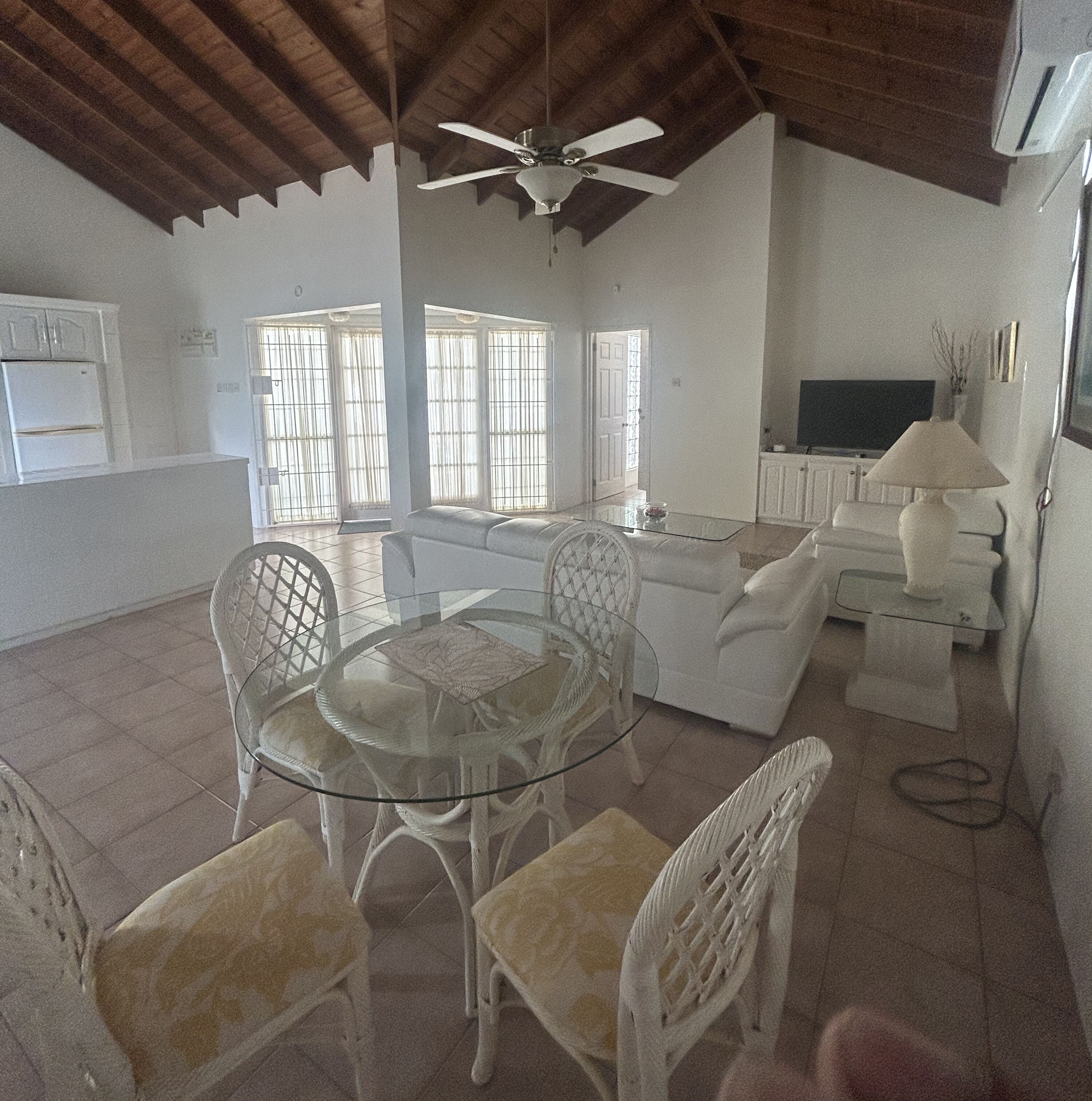 A spacious living area with a glass dining table, white wicker chairs, a sofa, and a television.