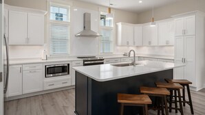 Une cuisine moderne avec des meubles blancs, un îlot central doté de tabourets, un four encastré et une hotte de cuisine en acier inoxydable.