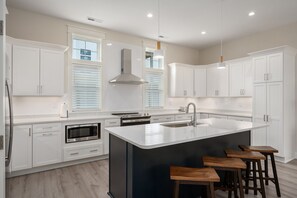 A modern kitchen with white cabinetry, a central island with stools, a built-in oven, and a stainless steel range hood. - Shorely Happy: New Construction, Semi-Oceanfront Townhouse, Close to Beach! (Kill Devil Hills)
