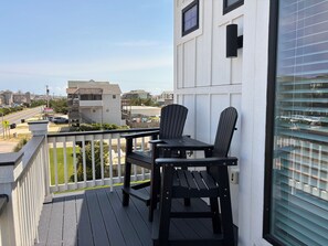 A balcony with a table and two chairs, overlooking a street with buildings and a clear sky. - Shorely Happy: New Construction, Semi-Oceanfront Townhouse, Close to Beach! (Kill Devil Hills)