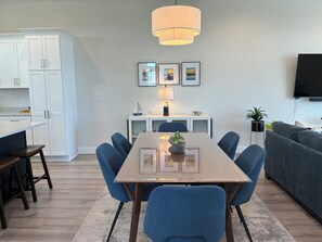 A modern living room with a dining table, blue chairs, a grey sofa, a flat-screen TV, and a kitchen area with white cabinets. - Shorely Happy: New Construction, Semi-Oceanfront Townhouse, Close to Beach! (Kill Devil Hills)