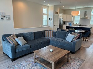 A modern living room with a sectional sofa, a coffee table, and a kitchen in the background. - Shorely Happy: New Construction, Semi-Oceanfront Townhouse, Close to Beach! (Kill Devil Hills)