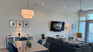 Un salon moderne comprenant un coin repas, un canapé gris, des chaises bleues, un téléviseur fixé au mur et un ventilateur de plafond.