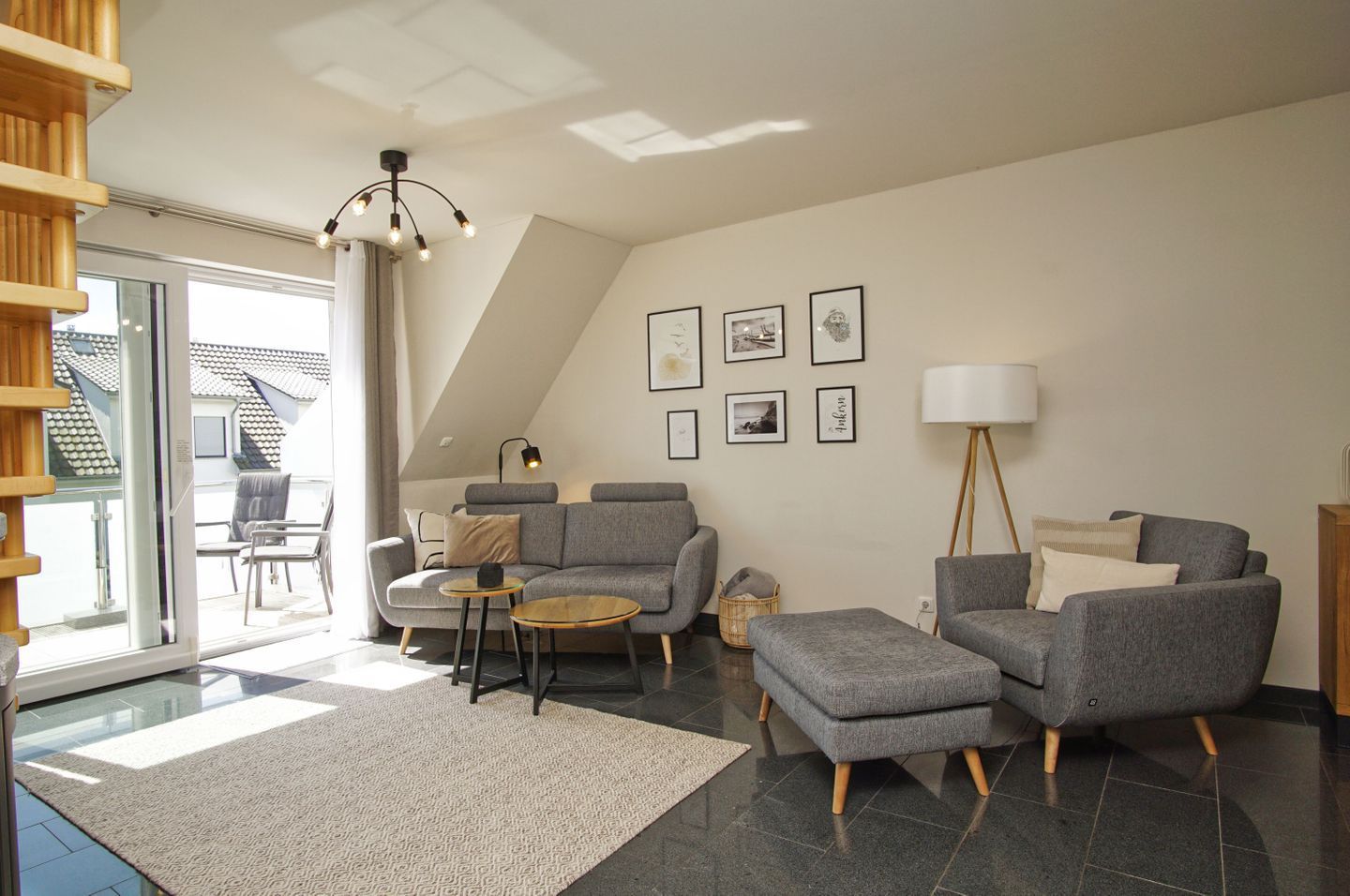 Een moderne woonkamer met een grijze zithoek, een salontafel en een vloerlamp. Er hangen ingelijste foto’s aan de muur en een trap leidt naar een buitenruimte.