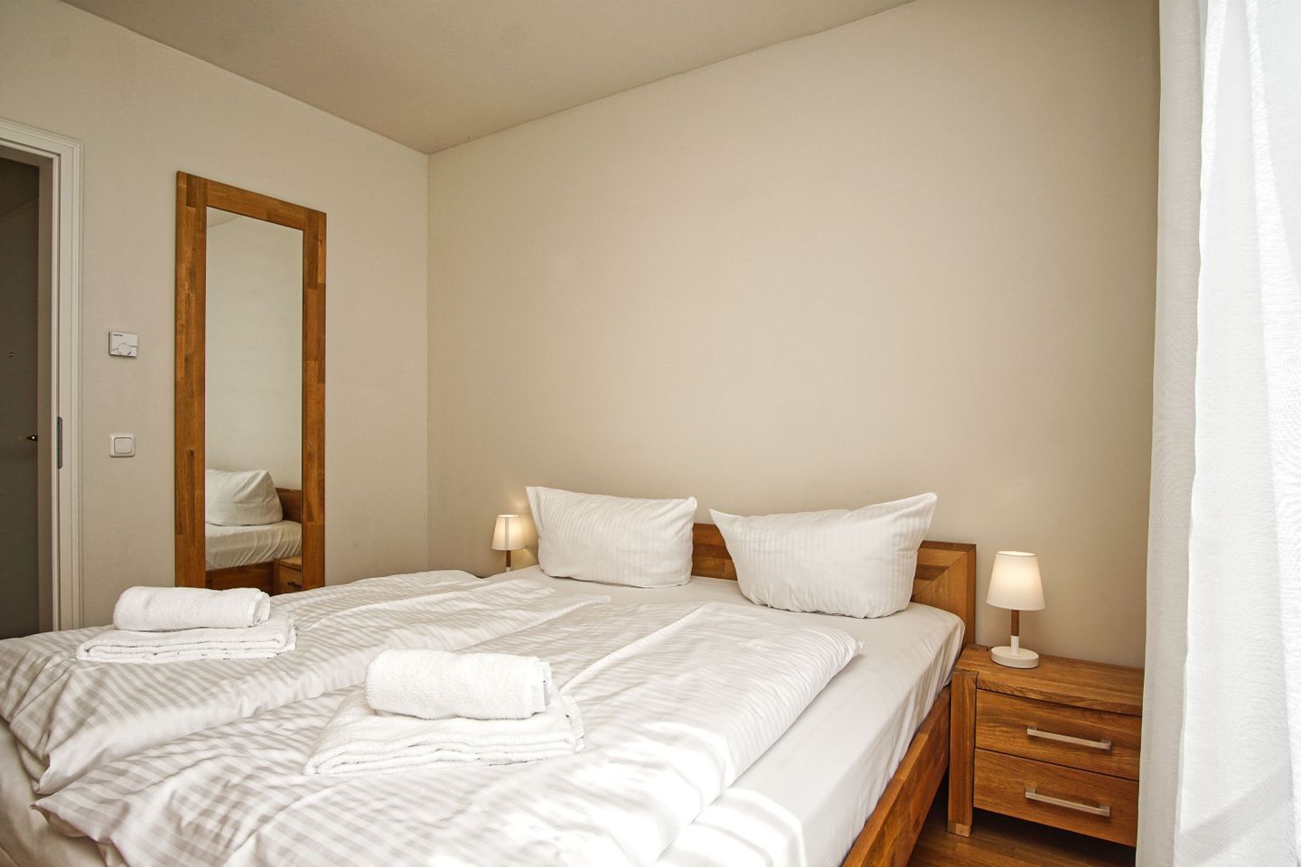 A neatly made bed with white linens and pillows, a wooden nightstand with a lamp, and a mirror on the wall.