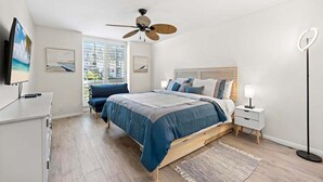 A bedroom with a bed, a ceiling fan, a bedside table, a floor lamp, and a window with a view. - Beautiful Upstairs Corner Cottage - Ocean view from Balcony NCV B2 (Oceanside)