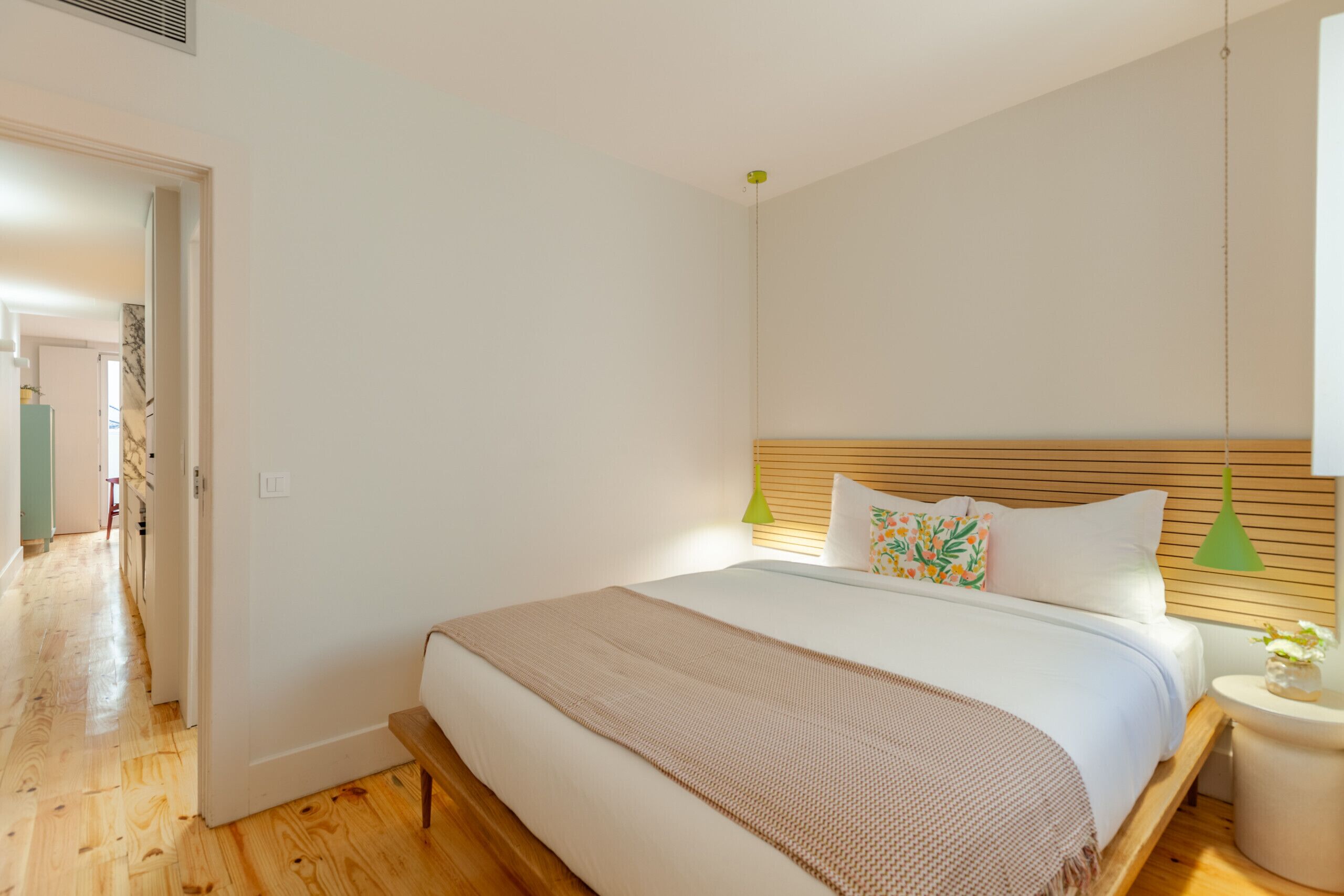 A bedroom with a wooden floor, a bed with a white headboard, two bedside lamps, and a small bedside table with a vase.