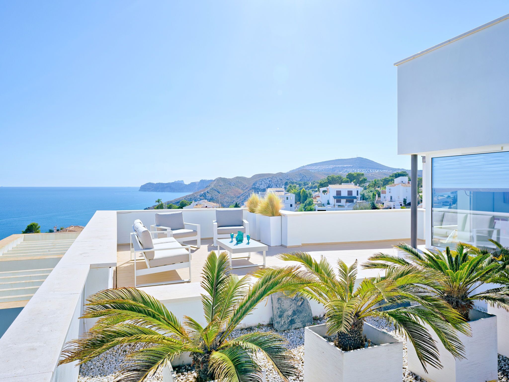 Une terrasse extérieure moderne, dotée de mobilier blanc et de palmiers, offrant une vue sur une ville côtière et la mer.