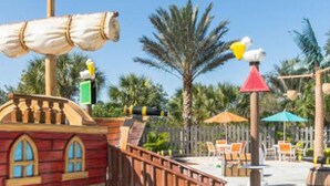 A wooden playhouse with a ship's wheel, palm trees, and outdoor seating.