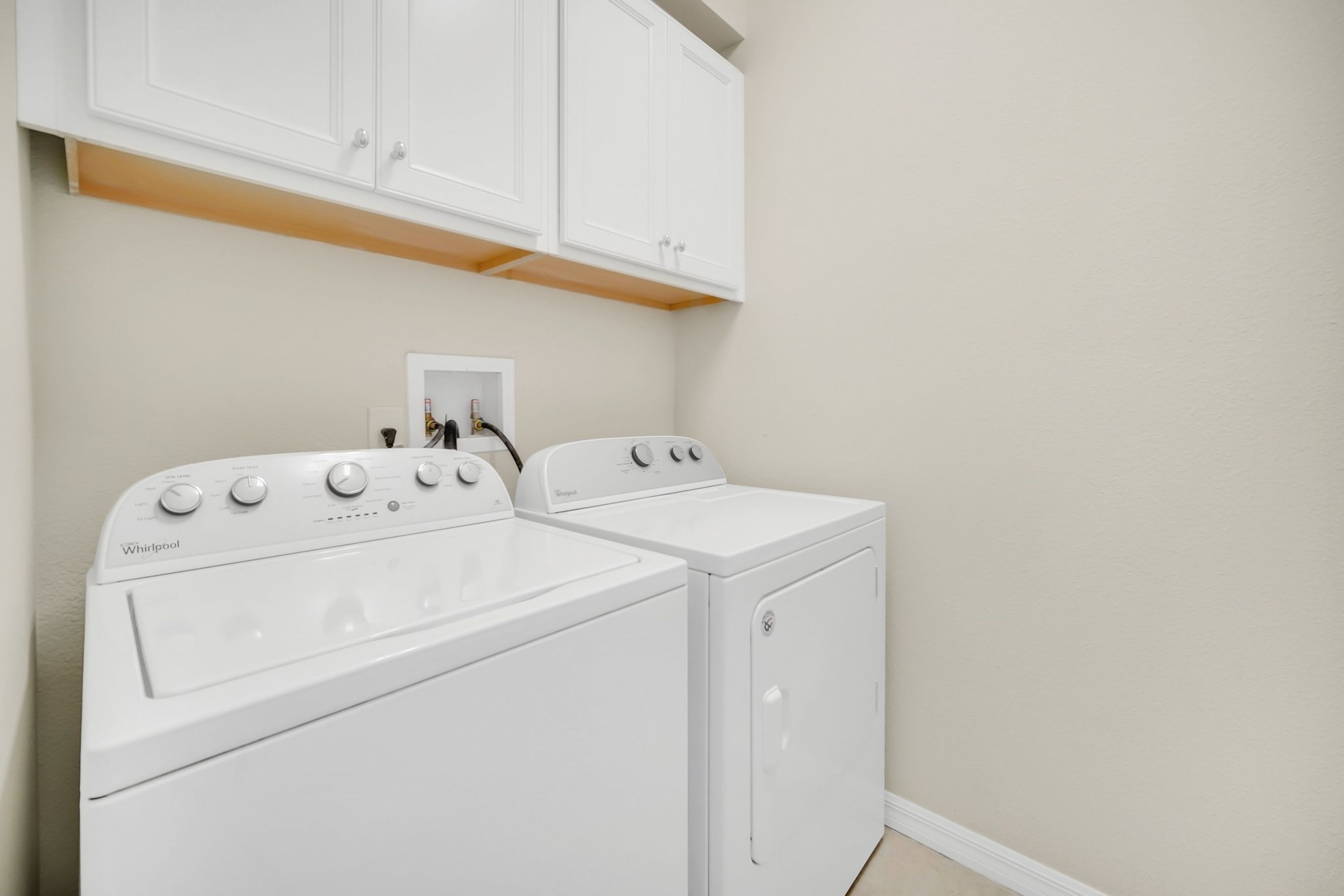 A laundry room with two white Whirlpool washers and dryers, white cabinets above, and a power outlet on the wall.
