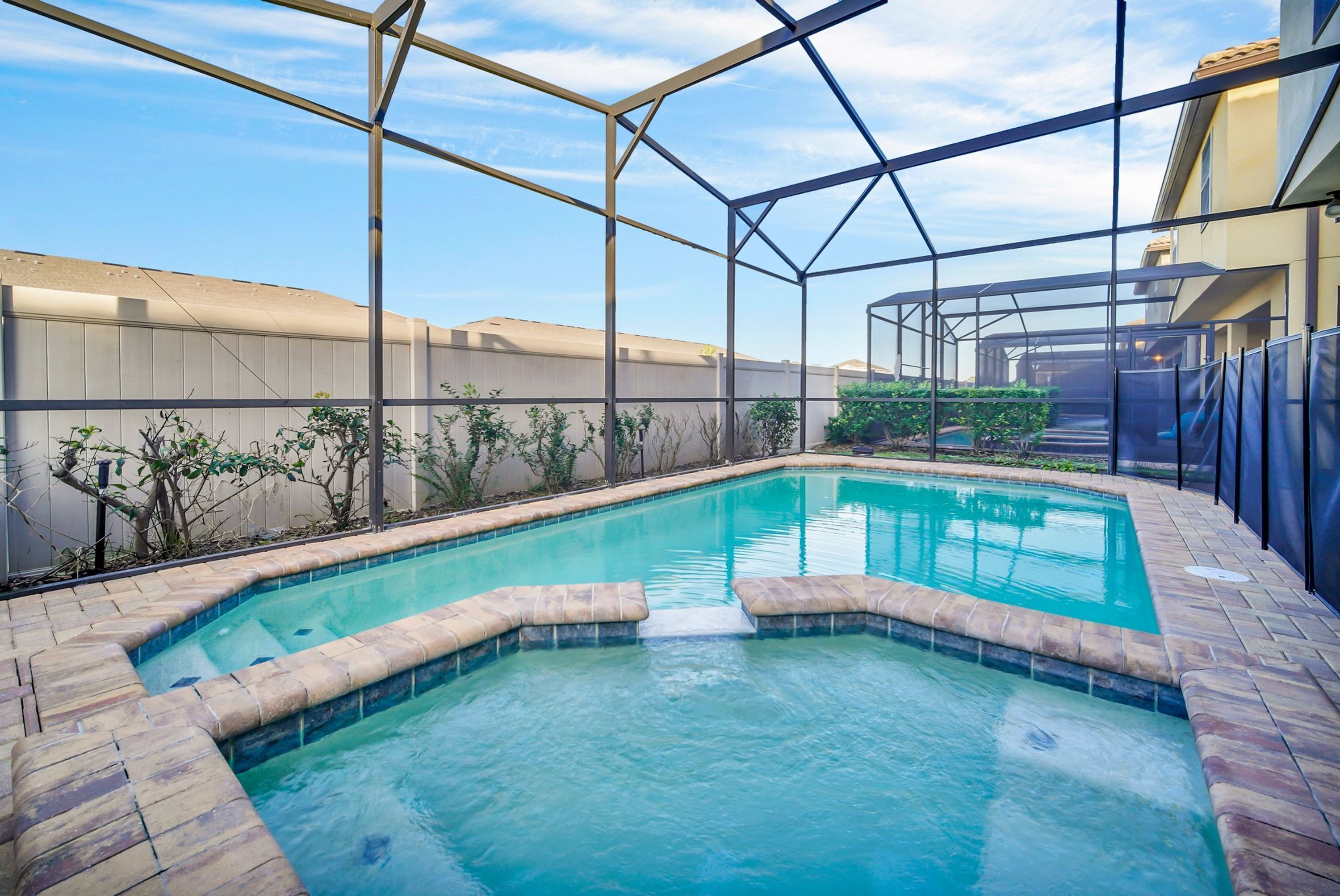 A swimming pool with a clear blue water, surrounded by a brick deck and a metal fence.