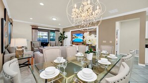 A dining area with a glass table set for six, surrounded by upholstered chairs. A chandelier hangs above the table, and a television is visible in the background.