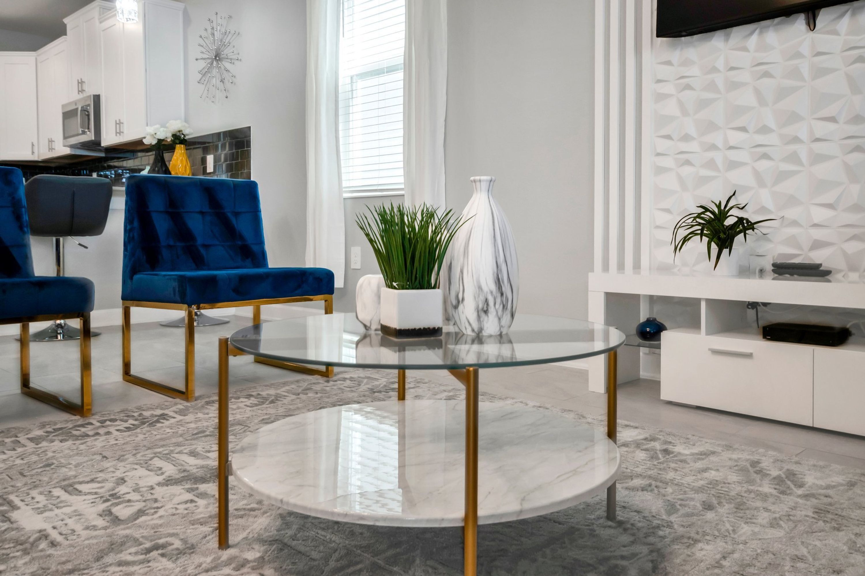 Uma sala de estar moderna com uma mesa de centro de vidro, poltronas de veludo azul, um vaso branco e um console branco com objetos decorativos.